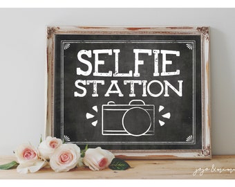 May include: A black chalkboard sign with white text that reads "SELFIE STATION" and a white silhouette of a camera. The sign is in a distressed white frame and is sitting on a wooden surface with pink roses in front of it.