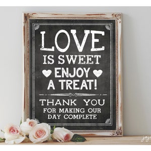 May include: A white framed chalkboard sign with the text "LOVE IS SWEET ENJOY A TREAT! THANK YOU FOR MAKING OUR DAY COMPLETE" in white lettering. The sign is sitting on a wooden surface with pink roses in front of it.