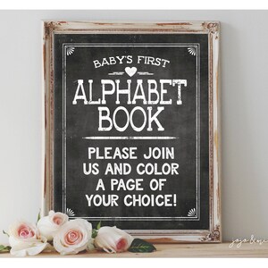 May include: A white sign with black lettering on a distressed wood frame. The sign says "Baby's First Alphabet Book" and "Please join us and color a page of your choice!"