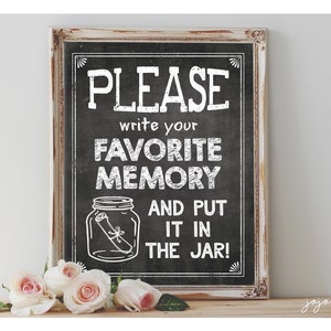 May include: A white sign with black lettering on a chalkboard background. The sign says "PLEASE write your FAVORITE MEMORY AND PUT IT IN THE JAR!" with a drawing of a jar with a rolled up piece of paper inside. The sign is in a white frame and is sitting on a wooden surface with a bouquet of white and pink roses in front of it.