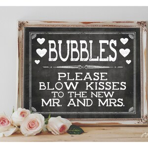 Op de afbeelding: Een zwart schoolbord met witte tekst die "Bubbles Please Blow Kisses To The New Mr. And Mrs." luidt. Het bord is ingelijst in een wit verweerd houten frame en staat op een houten oppervlak met witte rozen ervoor.