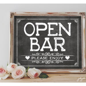 Puede incluir: Un letrero negro de pizarra con texto blanco que dice "Open Bar Please Enjoy" con dos corazones blancos a cada lado del texto. El letrero está enmarcado en un marco blanco y marrón y está sentado sobre una superficie de madera con rosas rosadas delante.