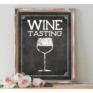 May include: A white sign with black lettering that says "Wine Tasting" with a white silhouette of a wine glass on a black background. The sign is framed in a white wooden frame.