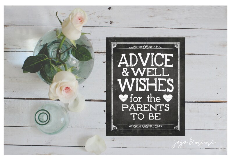 May include: A black chalkboard sign with white text that reads "Advice & Well Wishes for the Parents to Be". The sign is decorated with hearts and a decorative border. The sign is sitting on a white wooden surface with a vase of white roses.