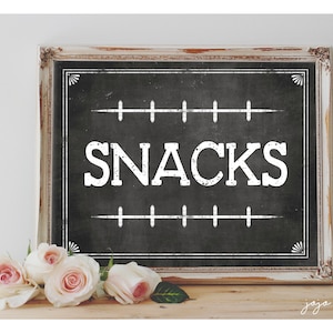May include: A white framed chalkboard sign with the word "SNACKS" written in white on a black background. The sign is sitting on a wooden surface with white roses in front of it.