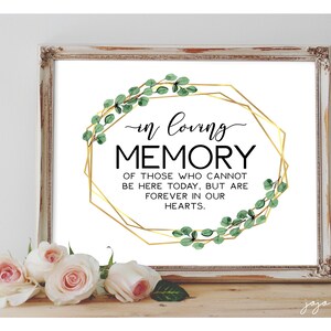 May include: A white framed sign with a gold geometric frame and green leaves around the frame. The sign says "in loving memory of those who cannot be here today, but are forever in our hearts."