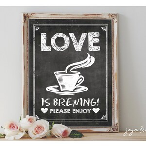 Op de afbeelding: Een wit gelijst bord van leisteen met de tekst "LOVE IS BREWING! PLEASE ENJOY" in witte krijt. Een witte koffiekop met stoom die eruit opstijgt bevindt zich in het midden van het bord. Het bord staat op een houten oppervlak met roze rozen ervoor.