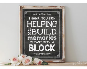 Puede incluir: Un cartel blanco con letras negras en un marco de madera rústico. El cartel dice "Gracias por ayudarnos a construir recuerdos. Por favor, firma un bloque." El cartel está sobre una superficie de madera con rosas rosas delante.
