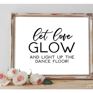 May include: White framed sign with black text that reads "Let love glow and light up the dance floor!" The sign is sitting on a wooden surface with white roses in front of it.