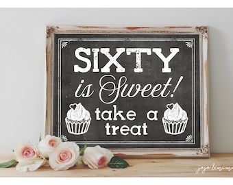 以下が含まれることがあります： 白い枠で囲まれた黒板の看板に「Sixty is Sweet! take a treat」という文字と、カップケーキのイラストが2つ描かれている。看板は木製の台の上に置かれ、その前に白いバラが置かれている。