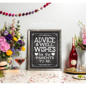 May include: A chalkboard sign with the text "Advice & Well Wishes for the Parents to Be" surrounded by flowers, a bottle of wine, and a glass of champagne. The sign is framed in a silver frame and has a black background with white text.