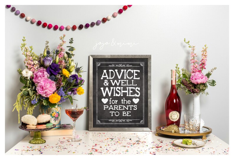 May include: A chalkboard sign with the text "Advice & Well Wishes for the Parents to Be" surrounded by flowers, a bottle of wine, and a glass of champagne. The sign is framed in a silver frame and has a black background with white text.