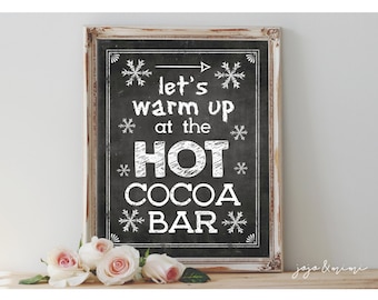May include: A framed chalkboard sign with the text "let's warm up at the HOT COCOA BAR" in white lettering. The sign is decorated with snowflake accents and a distressed white frame. Pink roses are in the foreground.