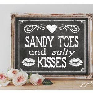 Puede incluir: Un letrero de pizarra negra con texto blanco que dice "Sandy Toes and Salty Kisses". El letrero está enmarcado en un marco blanco desgastado y está sentado sobre una superficie de madera. Hay rosas blancas en primer plano.