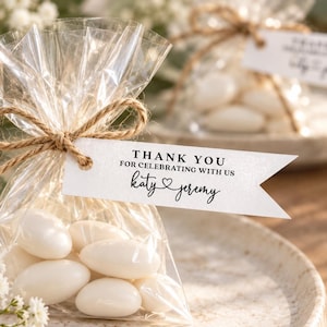 May include: A clear plastic favour bag filled with white almond sweets, tied with a brown twine bow. A white paper tag is attached, featuring the black printed text: THANK YOU FOR CELEBRATING WITH US, followed by the names Katy and Jeremy.