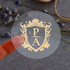 Op de afbeelding: Een heldere, ronde sticker met een gouden wapenontwerp. Het wapen toont de letters "P" en "A" gescheiden door een horizontale lijn. Bloemen en decoratieve elementen omringen de letters. De sticker is gedeeltelijk transparant.