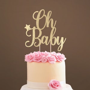 A cake topper made of gold glittered cardstock, spelling Oh Baby, decorated with butterflies. The font is hand-lettered typography.