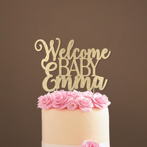 Personalized cake topper made of gold glittered cardstock, spelling Welcome BABY Emma. The font combines hand-lettered typography and a stylish script.