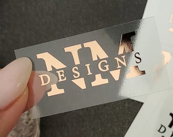 May include: A sheet of clear adhesive labels with the text "NM Designs" printed in rose gold foil. The labels are arranged in rows and columns on a white background.
