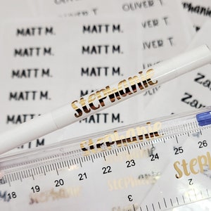 May include: A close-up of a white pen with gold lettering that says "Stephanie". The pen is lying on top of a clear ruler with black markings. The ruler is on top of a sheet of clear stickers with gold lettering. The stickers have the names "Zara", "Matt M.", "Stephanie", and "Oliver T" printed on them.