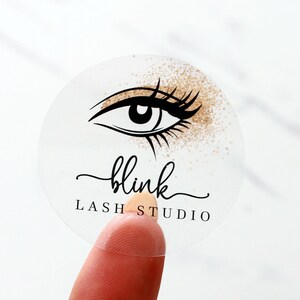 Può includere: Adesivo rotondo trasparente con un'illustrazione in bianco e nero di un occhio con glitter dorati. Il testo "blink lash studio" è stampato in nero sotto l'occhio.