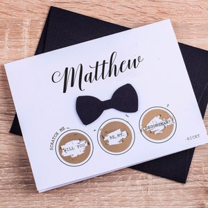 May include: A white greeting card featuring the name Matthew in black script, a black bow tie, and three circular scratch-off areas with the text Scratch Me, Will You, Be My, and Groomsman, signed by Ricky, placed on a black envelope.