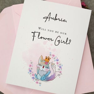 May include: A white card with pink watercolour accents and the text "Aubrie, Will you be our Flower Girl?"  A cartoon fox wearing a crown and surrounded by flowers is in the bottom centre of the card.