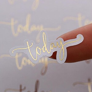 Peut inclure: Une feuille d'autocollants en feuille d'or avec le mot "aujourd'hui" écrit en cursive. Les autocollants sont sur un fond blanc.