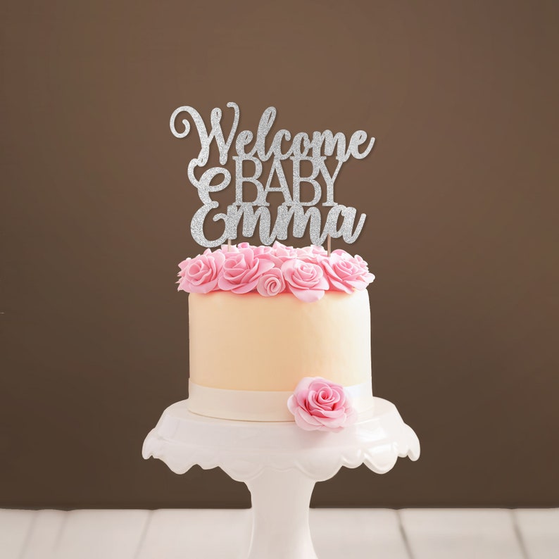 Personalized cake topper made of silver glittered cardstock, spelling Welcome BABY Emma. The font combines hand-lettered typography and a stylish script.