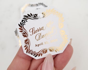 May include: White wedding favor tag with gold foil accents. The tag features a decorative border with a floral design. The text on the tag reads "Sierra & Daniel April 22, 2023".