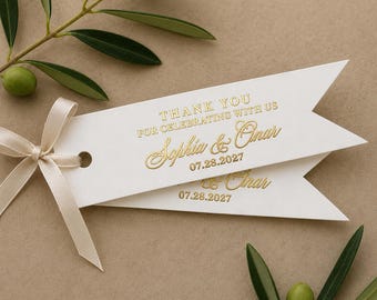 May include: Two white gift tags with gold text that reads "THANK YOU FOR CELEBRATING WITH US" and the names "Sophia & Onar" with the date "07.28.2027". A cream-colored ribbon is tied to the top tag. Olive branches with green olives are in the background.