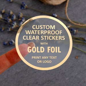 Puede incluir: Una pegatina circular transparente con un borde de lámina dorada y texto dorado que dice CUSTOM WATERPROOF CLEAR STICKERS WITH GOLD FOIL PRINT ANY TEXT OR LOGO. La pegatina tiene una superficie lisa sobre un fondo gris oscuro.