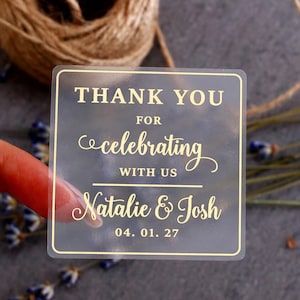 Può includere: Un adesivo quadrato semitrasparente con bordo dorato e testo dorato che recita: THANK YOU FOR celebrating WITH US Natalie & Josh 04. 01. 27. L'adesivo presenta una combinazione di caratteri in stampatello e corsivo centrati nel riquadro.