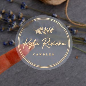 Könnte beinhalten: Ein runder, durchscheinender Aufkleber mit goldfarbenem Text und Grafiken. Das Design umfasst einen doppelten Linienrand, ein Blattzweig-Symbol, den Namen Kyla Riviera in Schreibschrift und das Wort CANDLES in einer einfachen, serifenlosen Schrift.
