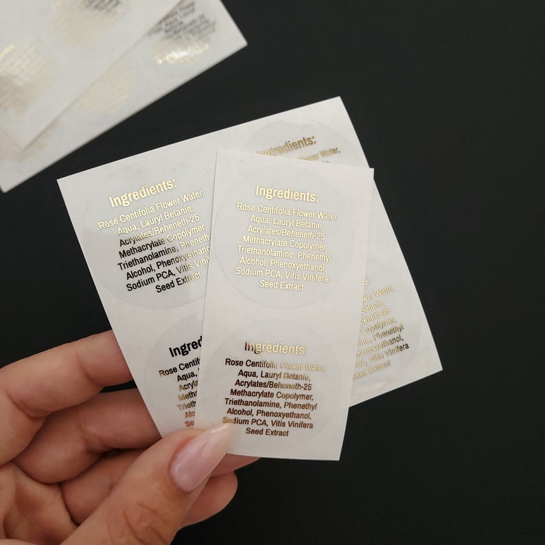 May include: Close-up of three round, white labels with gold lettering. The labels list the ingredients for a product, including Rose Centifolia Flower Water, Aqua, Lauryl Betaine, Acrylates/Beheneth-25 Methacrylate Copolymer, Triethanolamine, Phenethyl Alcohol, Phenoxyethanol, Sodium PCA, Vitis Vinifera Seed Extract.