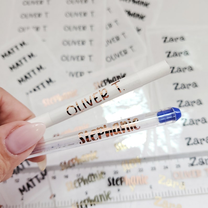 May include: Two clear pens with gold lettering. The pen on top has "Oliver T." written on it. The pen on the bottom has "Stephanie" written on it. The pens are on top of a sheet of paper with gold lettering that says "Zara" and "Oliver T."