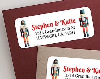 May include: A rectangular white address label featuring a nutcracker illustration on each side and the text Stephen & Katie, 1314 Grandheaven St, HAYWARD, CA 94541 printed in red and black. The label has rounded corners and a festive design.