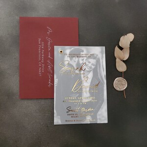 May include: A burgundy envelope with white calligraphy address and a rectangular translucent wedding invitation featuring a black and white couple photo, gold foil text, and a gold eyelet. A dried eucalyptus sprig with a wax seal is included.