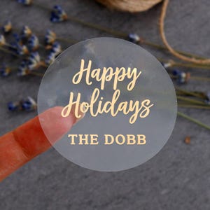 Può includere: Un adesivo circolare traslucido che presenta il testo color oro "Happy Holidays" in corsivo, seguito dal testo "THE DOBB" in un carattere grassetto senza grazie. L'adesivo ha una finitura liscia e semitrasparente.