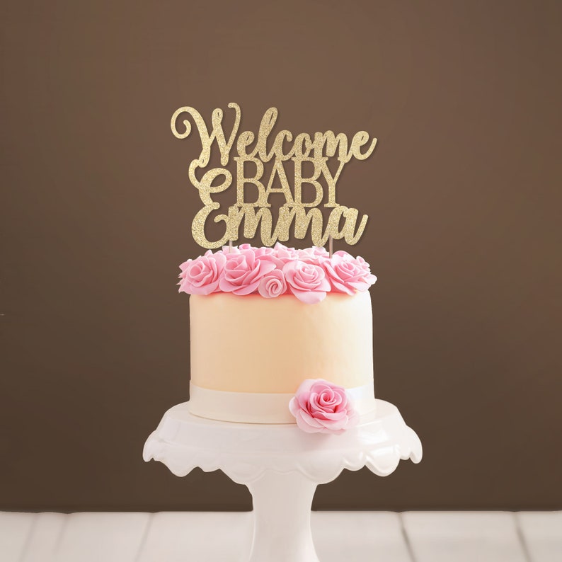 Personalized cake topper made of gold glittered cardstock, spelling Welcome BABY Emma. The font combines hand-lettered typography and a stylish script.