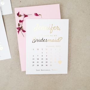 May include: A white card with gold foil text reading Jennifer, Will You Be My Bridesmaid? above an August 2023 calendar with a heart icon on the 26th and San Antonio, Texas at the bottom, placed on a light pink envelope with dried flower petals.