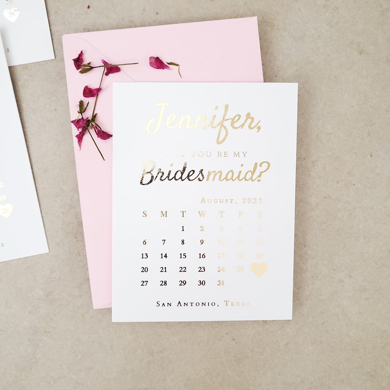 May include: A white card with gold foil text reading Jennifer, Will You Be My Bridesmaid? above an August 2023 calendar with a heart icon on the 26th and San Antonio, Texas at the bottom, placed on a light pink envelope with dried flower petals.