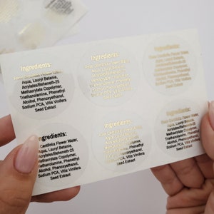 May include: Close-up of six round, clear labels with gold lettering. Each label reads "Ingredients:" followed by a list of ingredients for a skincare product. The ingredients include Rose Centifolia Flower Water, Aqua, Lauryl Betanie, Acrylates/Beheneth-25 Methacrylate Copolymer, Triethanolamine, Phenethyl Alcohol, Phenoxyethanol, Sodium PCA, and Vitis Vinifera Seed Extract.