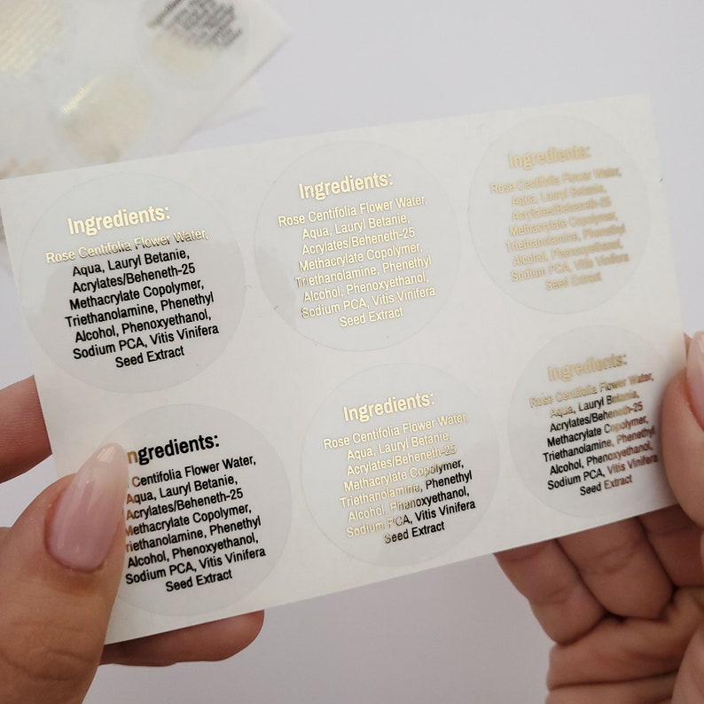 May include: Close-up of six round, clear labels with gold lettering. Each label reads "Ingredients:" followed by a list of ingredients for a skincare product. The ingredients include Rose Centifolia Flower Water, Aqua, Lauryl Betanie, Acrylates/Beheneth-25 Methacrylate Copolymer, Triethanolamine, Phenethyl Alcohol, Phenoxyethanol, Sodium PCA, and Vitis Vinifera Seed Extract.