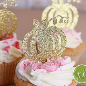 Peut inclure: Deux cupcakes surmont&eacute;s de gla&ccedil;age blanc et de p&eacute;pites roses. Chaque cupcake est d&eacute;cor&eacute; d'un topper en forme de citrouille dor&eacute;e &agrave; paillettes.