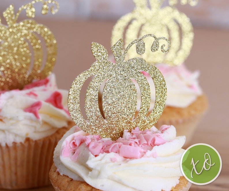 Peut inclure: Deux cupcakes surmont&eacute;s de gla&ccedil;age blanc et de p&eacute;pites roses. Chaque cupcake est d&eacute;cor&eacute; d'un topper en forme de citrouille dor&eacute;e &agrave; paillettes.