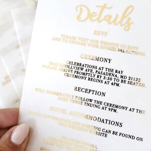 Op de afbeelding: Gouden bruiloftsuitnodigingskaart met een witte achtergrond. De kaart bevat informatie over de ceremonie, de receptie en de hotelaccommodatie. De tekst luidt: "Details", "RSVP", "Bezoek onze website om je aanwezigheid te bevestigen en je dinerkeuze te maken", "Ceremonie", "Vieringen aan de baai", "2043 Knollview Ave, Pasadena, MD 21122", "Wees er om 15:30 uur om plaats te nemen", "De ceremonie begint om 16:00 uur", "Receptie", "De receptie volgt direct na de ceremonie op dezelfde locatie en eindigt om 21:00 uur", "Hotelaccommodatie", "Informatie over het hotel en boekingen is te vinden op onze trouwwebsite", "Voor meer details en verlooffoto's, bezoek onze website."