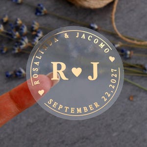 Puede incluir: Una pegatina redonda transparente con letras doradas. La pegatina muestra los nombres "Rosalinda & Jacomo" y la fecha "22 de septiembre de 2027". Las iniciales "R" y "J" están en el centro, con un símbolo de corazón entre ellas. También hay pequeños iconos de corazón.