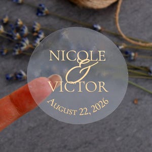 May include: A transparent, circular sticker with gold text. It reads "Nicole & Victor" with "August 22, 2026" below. The sticker is held by a finger, with dried lavender and twine in the background.