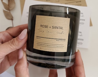 Può includere: Una candela in cera di soia in un barattolo di vetro nero con un'etichetta di carta marrone su cui è scritto "ROSE + SANTAL Soy Wax Candle" e "Made in the USA". L'etichetta include anche il testo "Infused with sensual woody ambery fragrance blend" e "MalovaCandles.com".